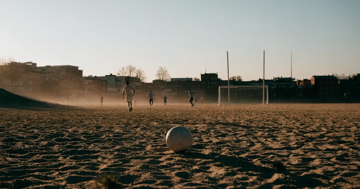 Ein sandiger Fußballplatz am Rand einer Stadt in warmem Abendlicht, Staub in der Luft und zwei Tore in der Ferne.