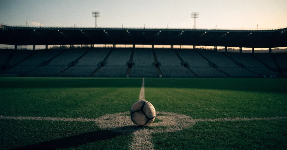 Ein Fußball liegt auf dem Anstoßpunkt eines leeren Rasenplatzes, im Hintergrund eine große Stadiontribüne im Abendlicht.