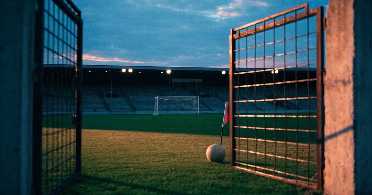 Blick durch ein Stadiontor auf einen grünen Rasen im Abendlicht, ein Ball ruht an der Eckfahne, die Tribünen sind leer.
