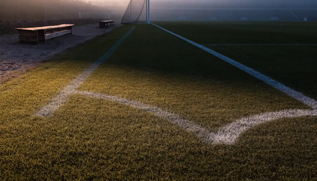 Ein Fußballplatz mit frisch gezogenen Kreidelinien im ersten Morgenlicht, leere Ersatzbänke am Spielfeldrand.
