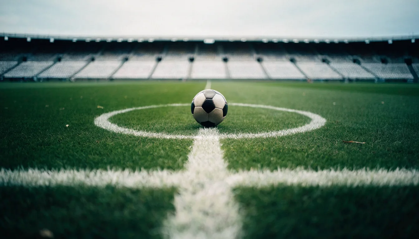 Ein Fußball liegt auf dem Rasen im Mittelkreis, die Kreidelinien führen scharf ins Bild, dahinter verschwimmt eine Tribüne.