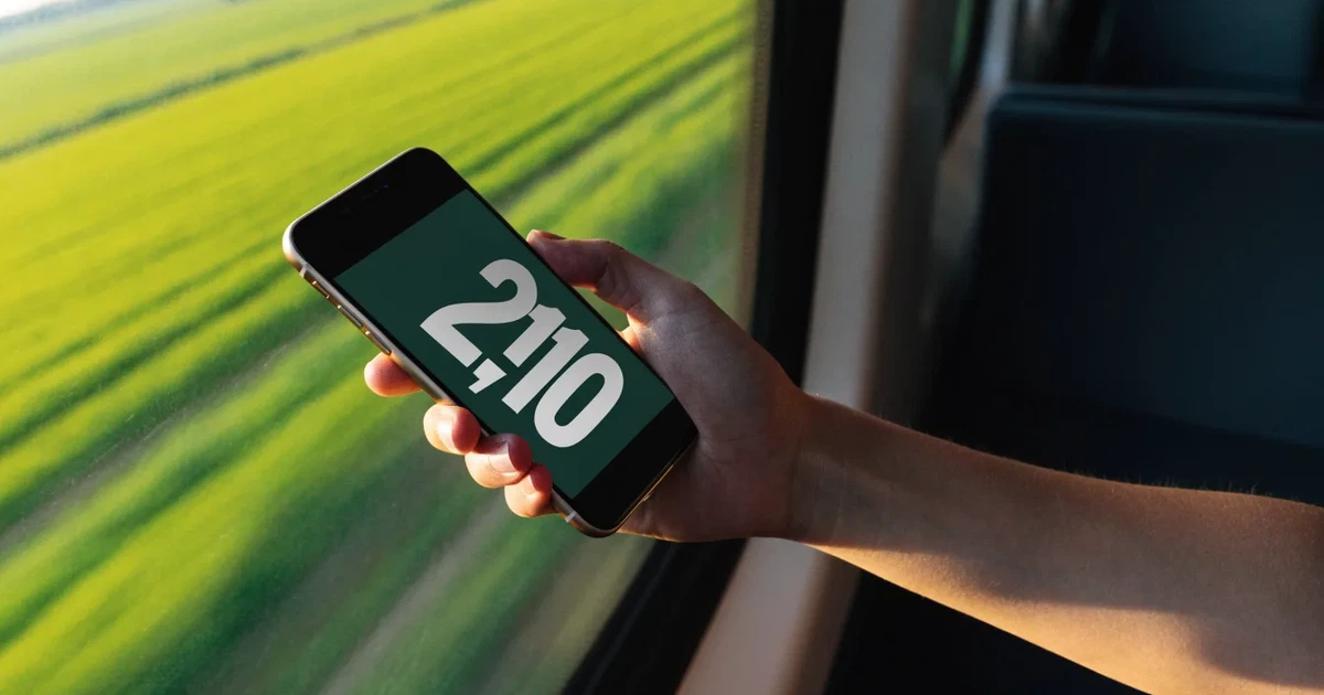 Eine Hand hält ein Smartphone mit dunklem Display in einem Zug, am Fenster ziehen verschwommen Felder vorbei.