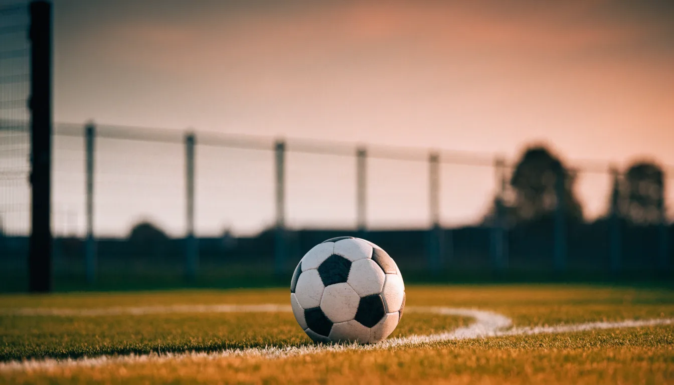 Ein Fußball liegt am Rand eines Rasens neben einer weißen Linie, im Hintergrund ein Zaun in der Abendsonne.