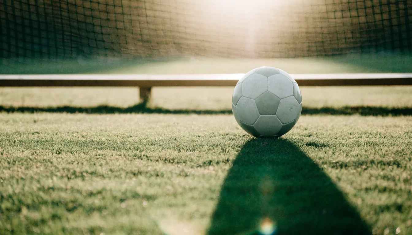 Ein Fußball auf einem Rasenplatz im Gegenlicht der tiefstehenden Sonne, am Rand eine leere Ersatzbank.