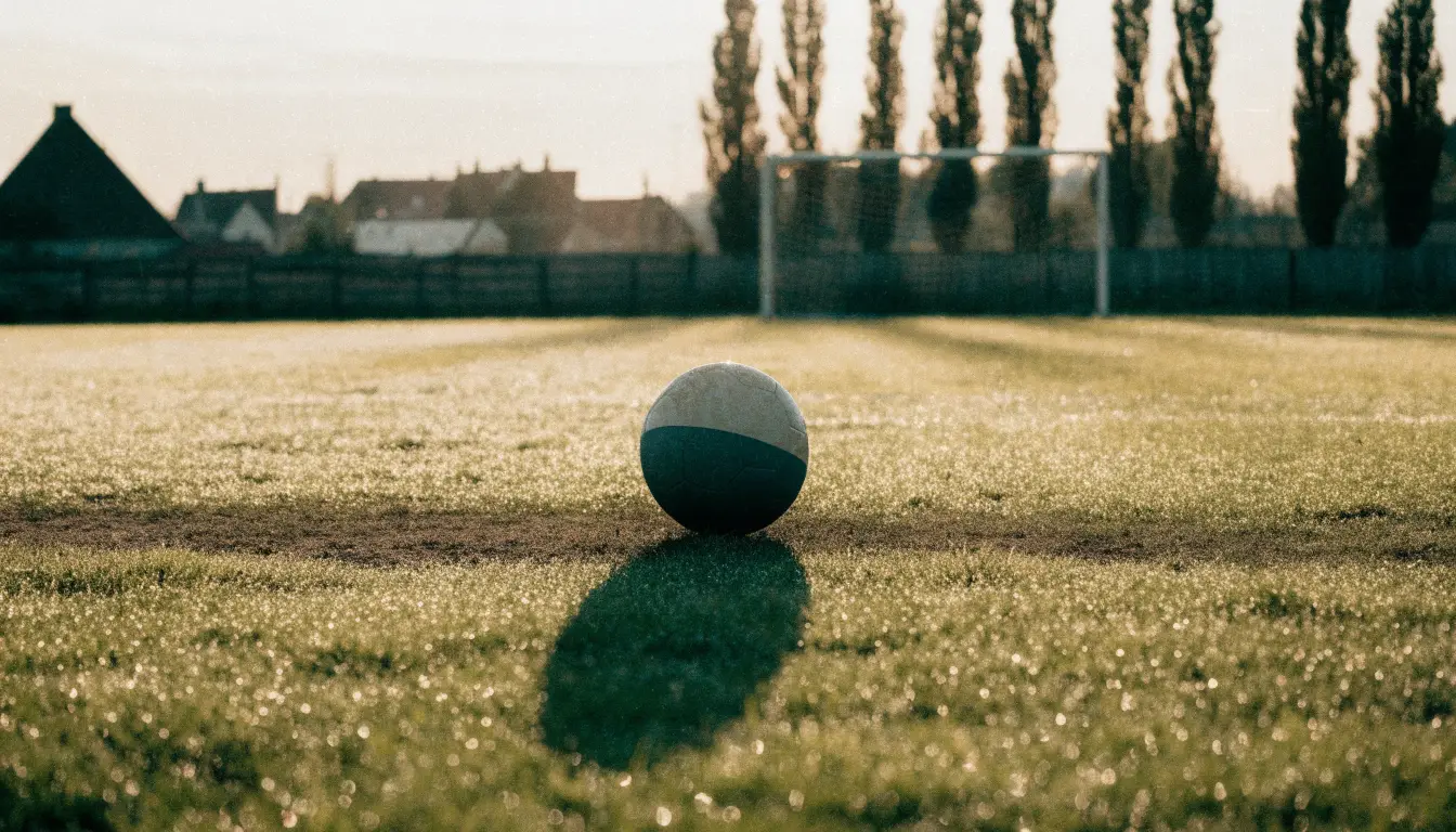 Ein Fußball auf dem Rasen eines leeren Sportplatzes am Rand einer Kleinstadt im Morgenlicht.