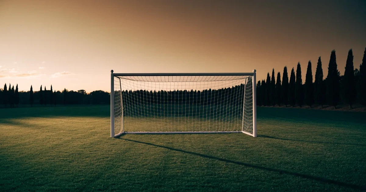 Ein Fußballtor mit Netz auf einem leeren Platz im Abendlicht, der Rasen frisch gemäht, im Hintergrund Zypressen.