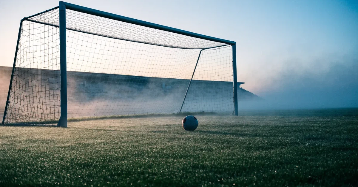 Ein Fußballtor mit Netz steht auf einem leeren Platz im Nebel der Morgendämmerung, der Rasen ist taufeucht.