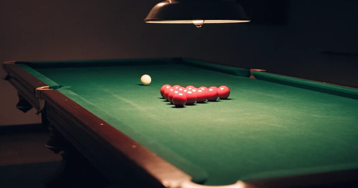 Ein Snookertisch mit grünem Tuch und aufgebauten roten Kugeln in einem abgedunkelten Raum unter einer hängenden Lampe.