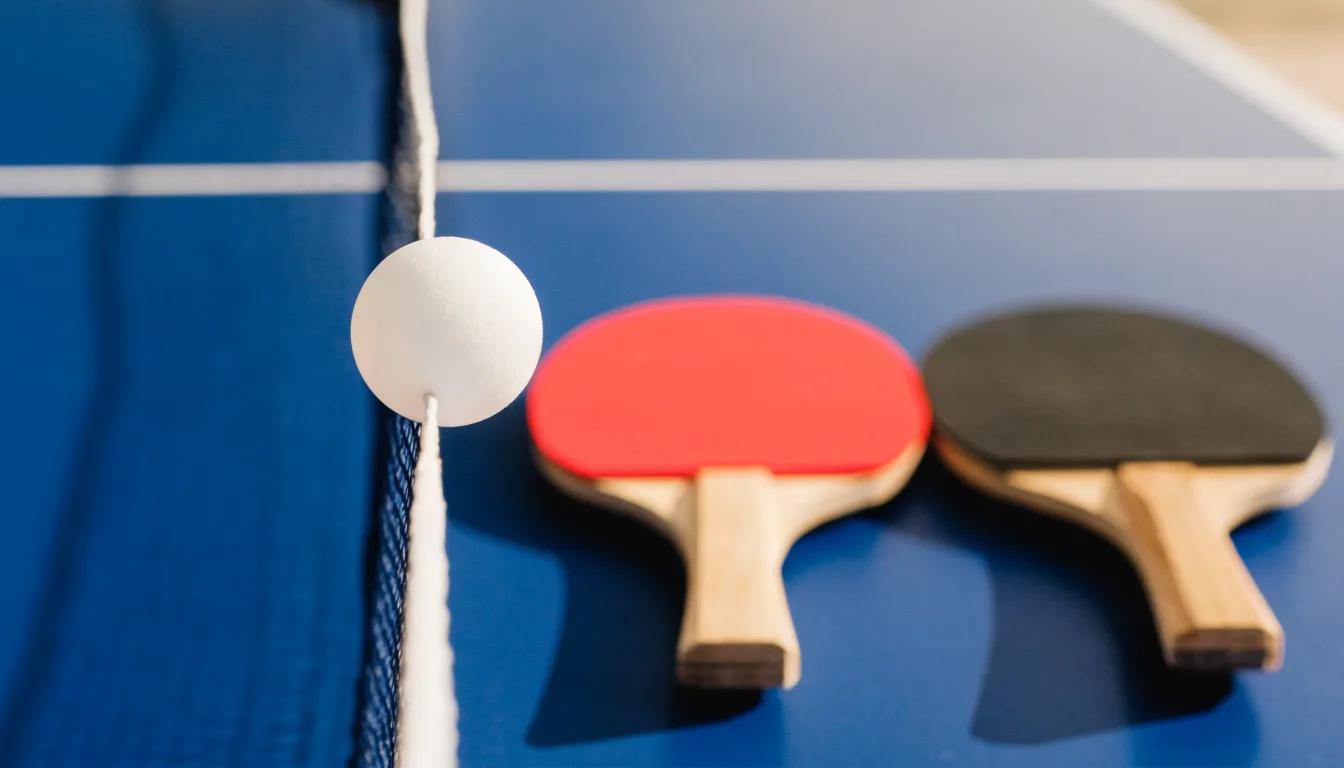 Ein weißer Ball liegt in Nahaufnahme auf der Netzkante einer Tischtennisplatte, daneben zwei Holzschläger mit rotem und schwarzem Belag.