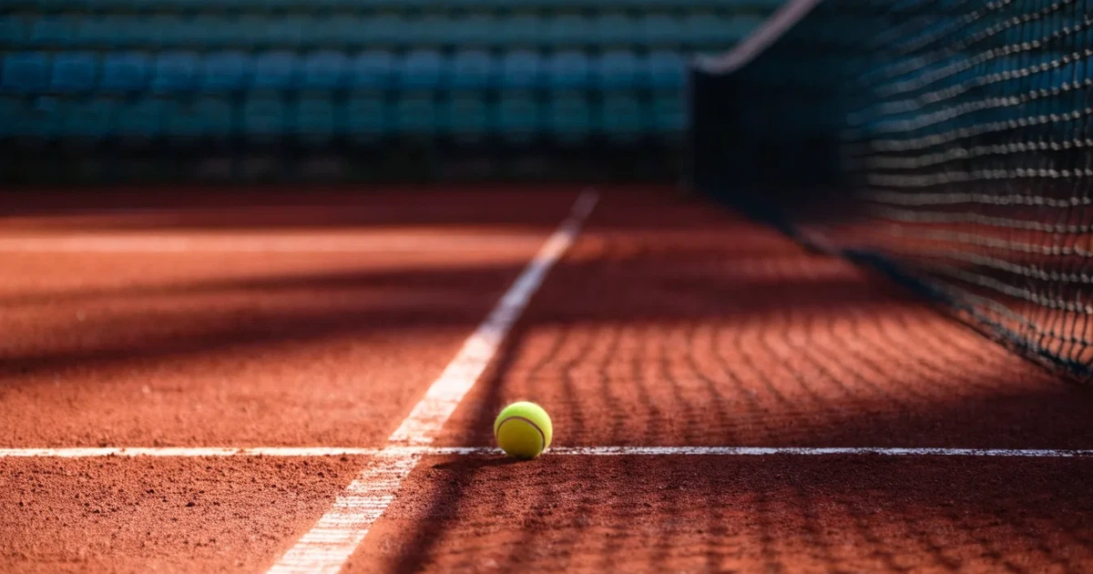 Ein Sandplatz mit rötlichem Belag und gezogenen weißen Linien, ein einzelner Tennisball liegt neben der Linie, im Hintergrund eine leere Tribüne.