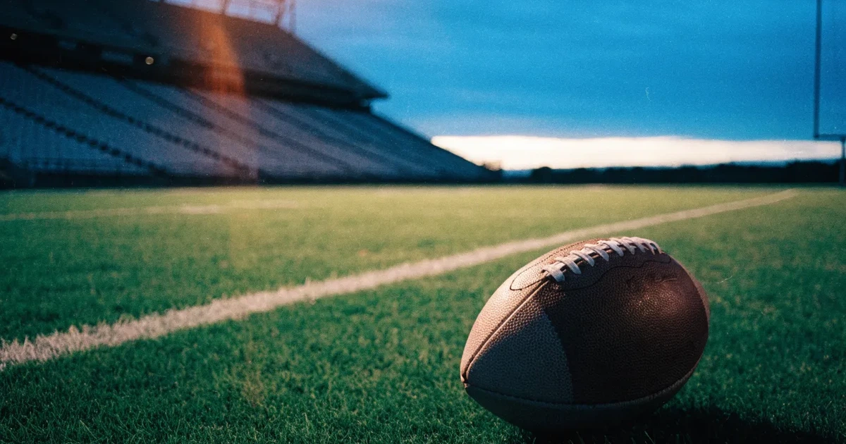 Ein American-Football-Ball liegt auf einem Rasenplatz unter Flutlicht, im Hintergrund eine große Tribüne in der Dämmerung.