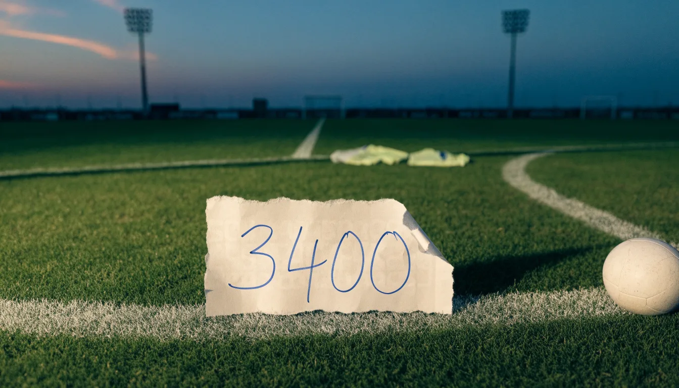 Ein Fußball liegt auf dem Anstoßpunkt eines Rasenplatzes in der Abenddämmerung, Flutlichtmasten stehen unscharf im Hintergrund.