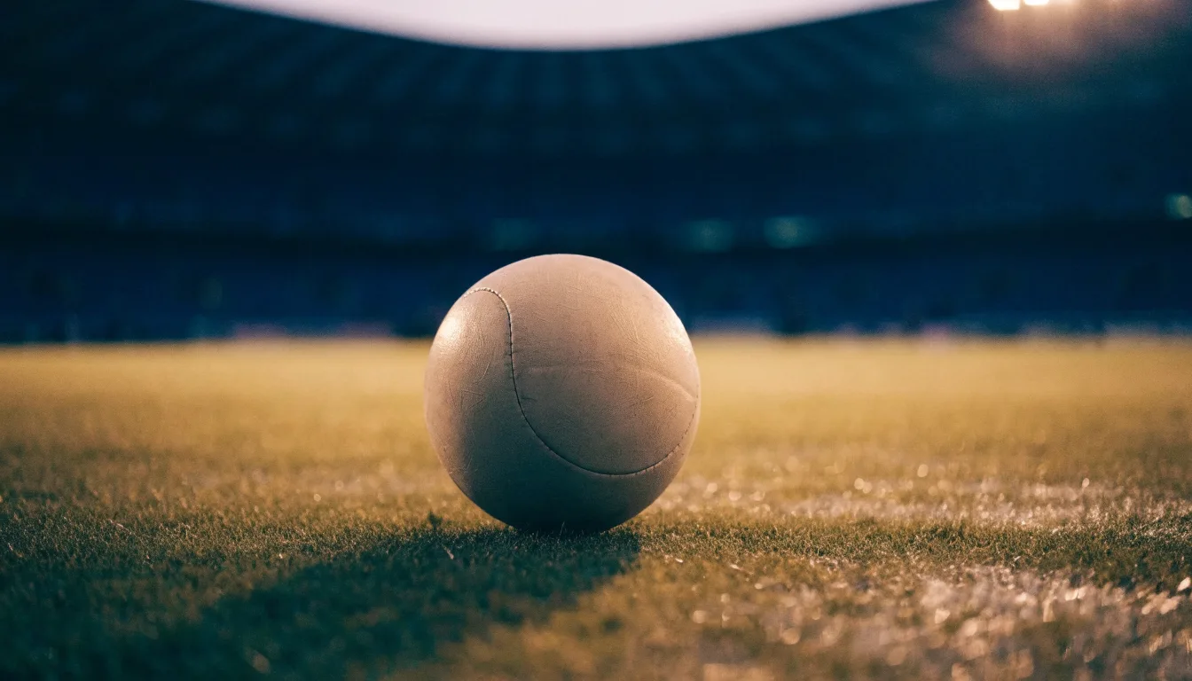 Ein Fußball liegt auf einem Rasenplatz in der Dämmerung, im Hintergrund ein ferner Stadionrand unter Flutlicht.